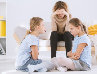 Young pouty sisters arguing with arms crossed and their stressed mom sitting on a sofa in the background